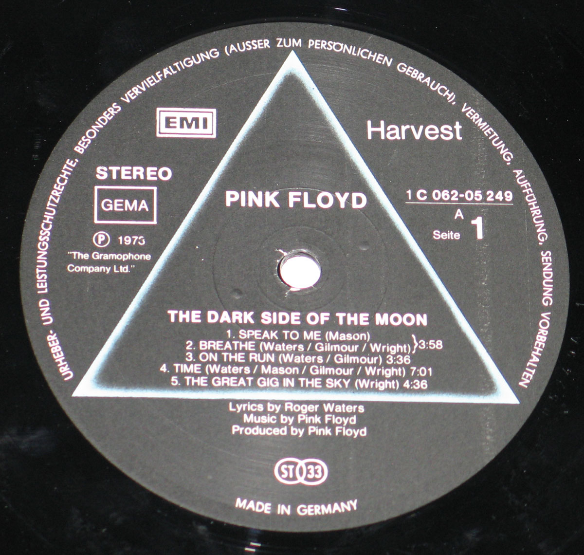 ‘Record label of Pink Floyd’s The Dark Side of the Moon, German pressing by EMI on Harvest label, catalog number 1C 062-05 249. Black label with a blue triangle prism design, featuring track listings for Side A and credits to Roger Waters, David Gilmour, Nick Mason, and Richard Wright. Includes “GEMA” and “ST 33” logos, with text “Made in Germany” at the bottom.’